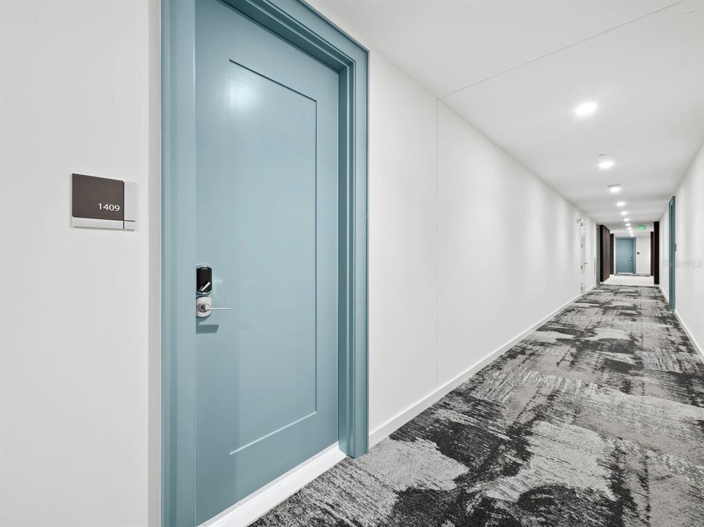 A light blue door with a silver handle stands in a long, carpeted apartment hallway — 301 QUAY COMMONS UNIT 1409, SARASOTA, FL