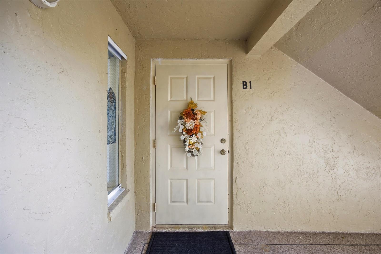 A beige front door with a floral wreath and the number B1 sits under an overhang — 722 Sunny Pine Way # B1, FL 33415, FL