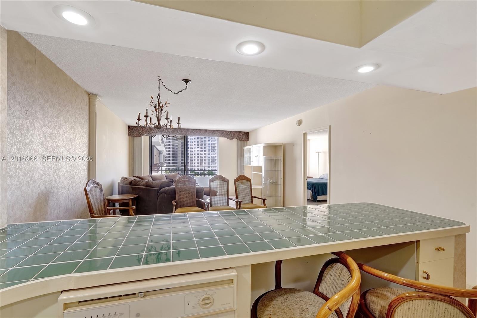 A kitchen counter with green tiles overlooks a living room with a chandelier and furniture — 500 Three Islands Blvd # 622, Hallandale Beach FL 33009, HALLANDALE BEACH, FL