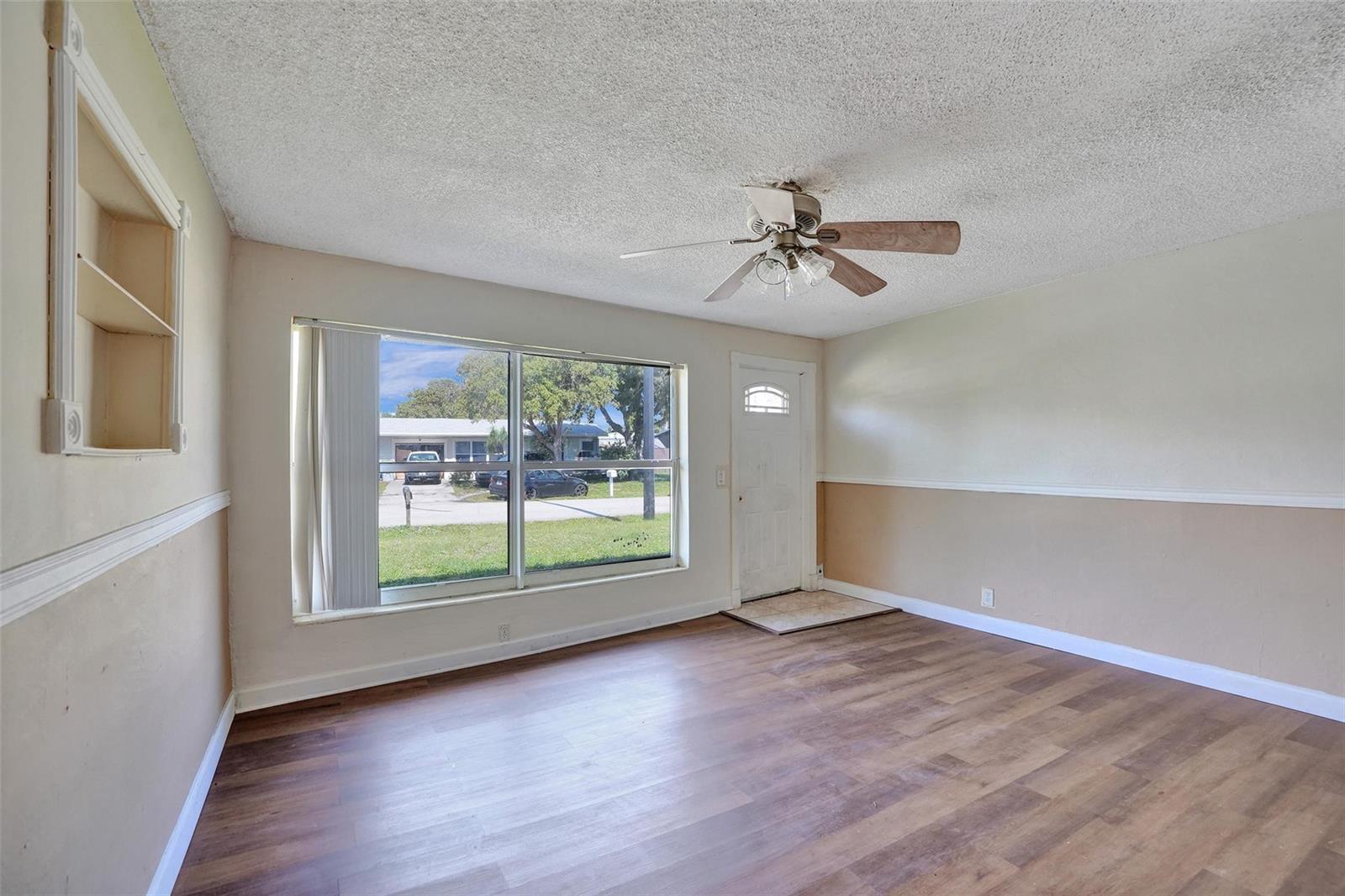 An empty living room features wood-look flooring, a ceiling fan, a large window, and wainscoting — 1560 NE 31st Court, Pompano Beach FL 33064, POMPANO BEACH, FL
