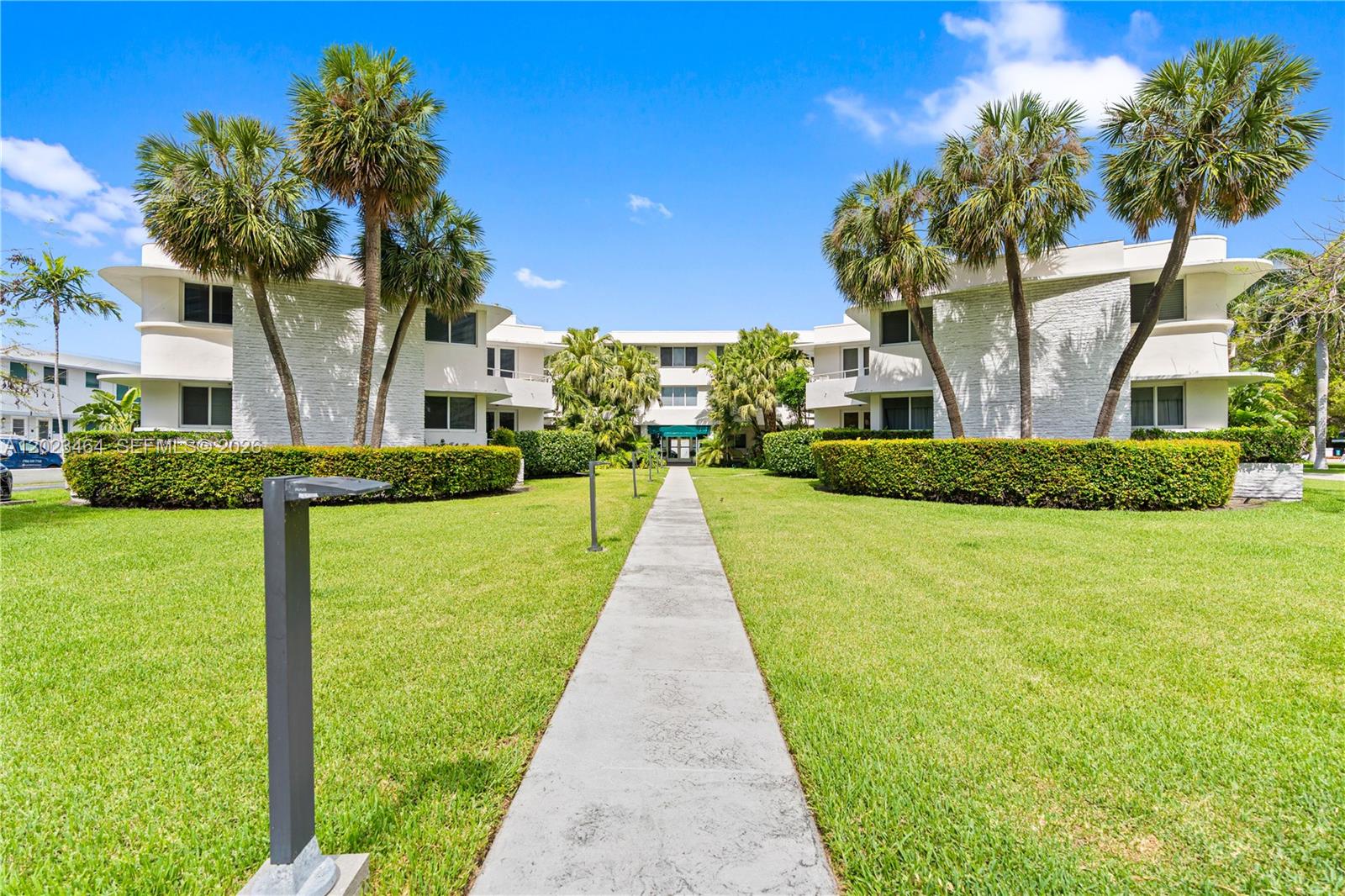 A concrete walkway leads through a grassy courtyard toward a multi-story white apartment building — 10190 Collins Ave, Bal Harbour FL 33154, BAL HARBOUR, FL