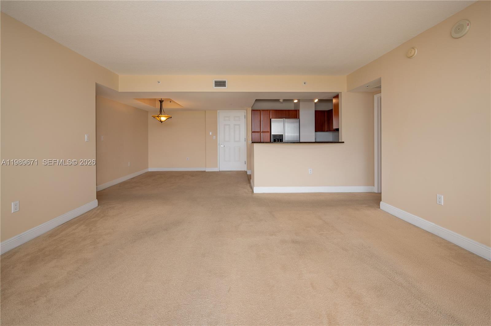 An empty living area with beige carpet leads to an open kitchen with wooden cabinets — 3232 SW Coral Way # 1504, Miami FL 33145, MIAMI, FL
