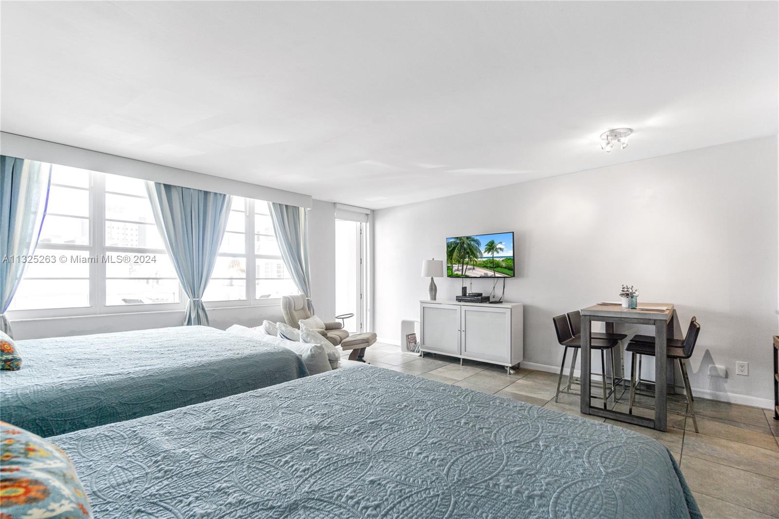 A bedroom features two beds, a dining table, a television, and large windows with curtains — 100 Lincoln Rd # 320, Miami Beach FL 33139, MIAMI BEACH, FL
