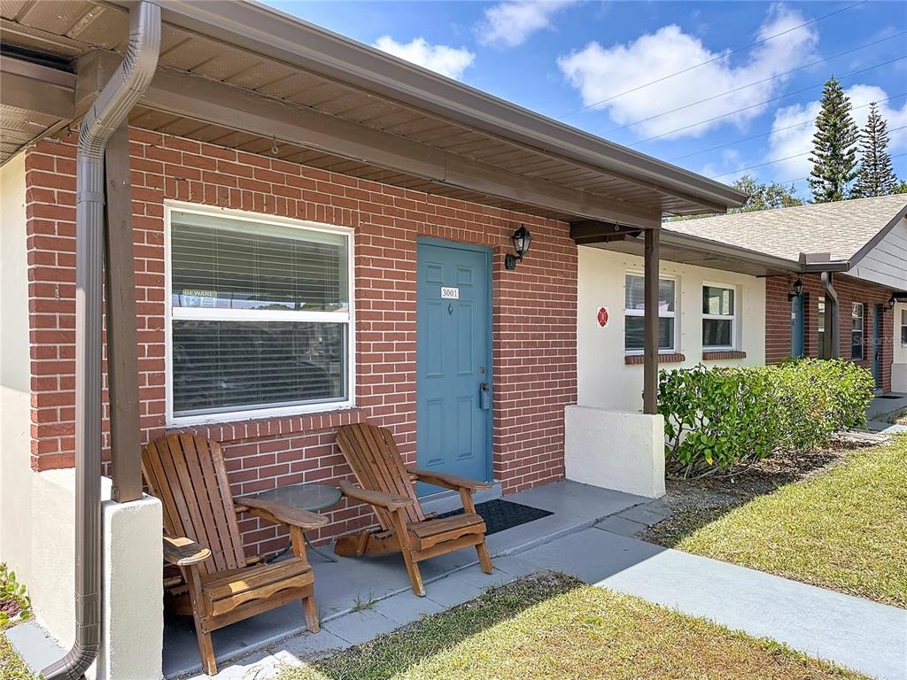 A brick exterior building features a blue front door, two wooden chairs, and a walkway — 24862 US HIGHWAY 19 N UNIT 3001, CLEARWATER, FL