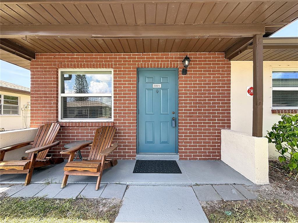 A brick exterior features a blue front door, a covered porch, and two wooden chairs — 24862 US HIGHWAY 19 N UNIT 3001, CLEARWATER, FL