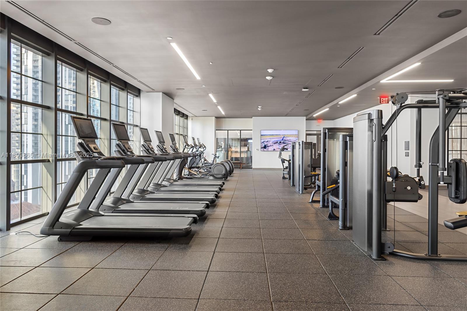 A modern gym features a row of treadmills, strength training equipment, and floor-to-ceiling windows — 159 NE 6th St # 3610, Miami FL 33132, MIAMI, FL