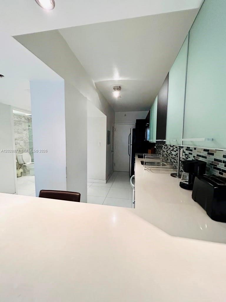 A kitchen with white countertops and a tiled backsplash opens to a hallway and bathroom — 100 Lincoln Rd # 701, Miami Beach FL 33139, MIAMI BEACH, FL