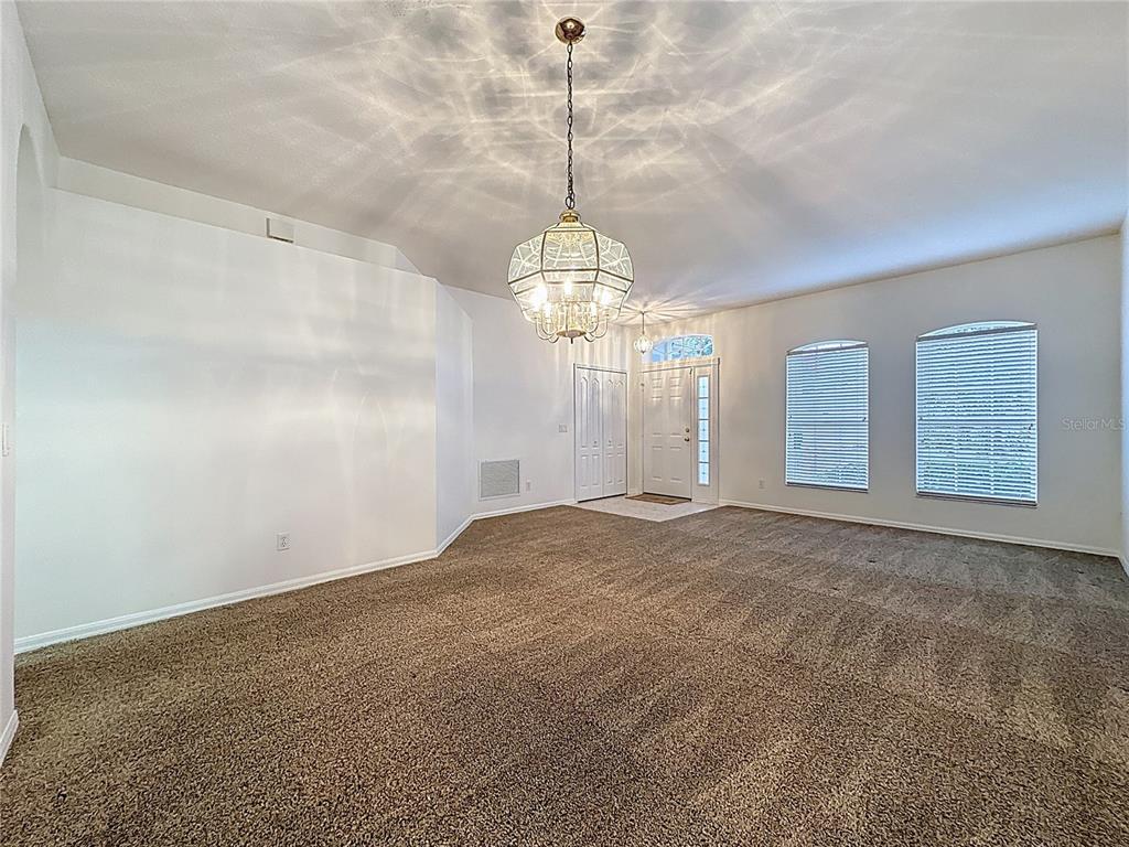 An empty living room features beige carpeting, white walls, two chandeliers, and a front door — 4246 NEWLAND STREET, CLERMONT, FL
