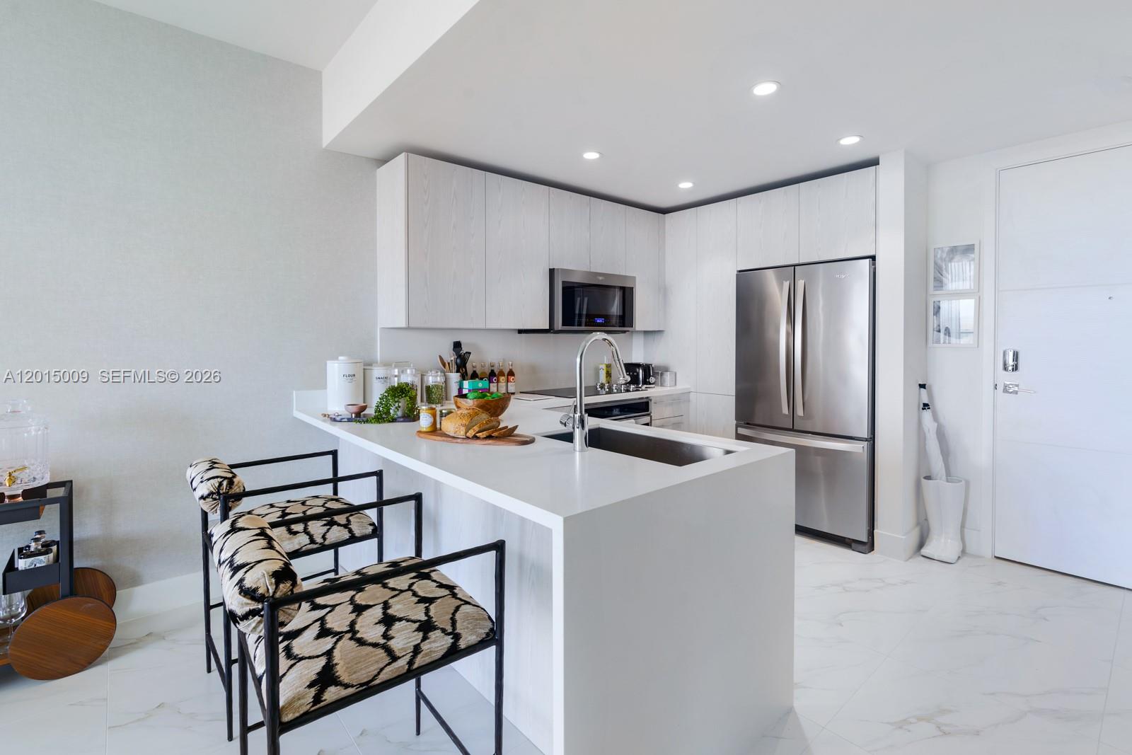 A modern kitchen features white cabinetry, a waterfall island with a sink, and patterned stools — 2000 Metropica Way # 1207, Sunrise FL 33323, SUNRISE, FL