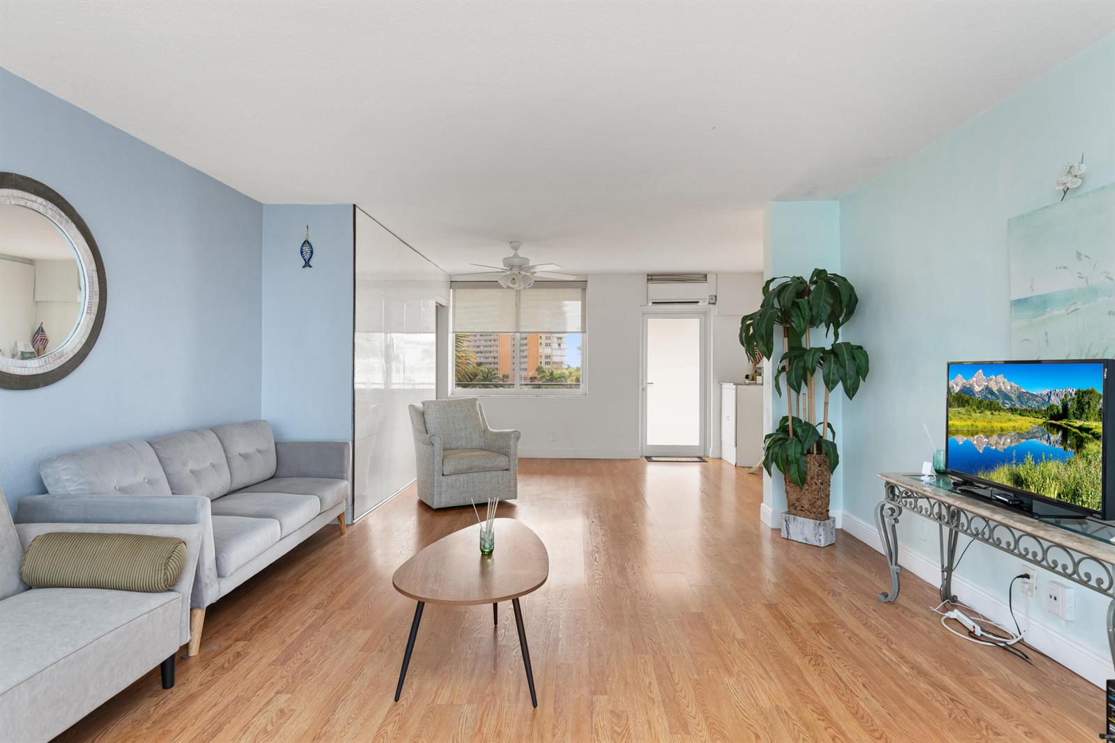 A living room features light blue walls, wood-look flooring, a gray sofa, and a television — 401 N Riverside Drive # 406, Pompano Beach FL 33062, POMPANO BEACH, FL