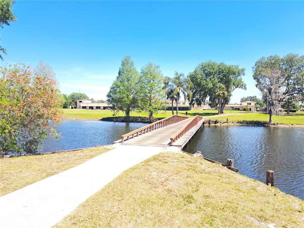 A wooden footbridge crosses a pond toward a grassy lawn and a distant building — 604 CAMELLIA DRIVE, INDIAN LAKE ESTATES, FL