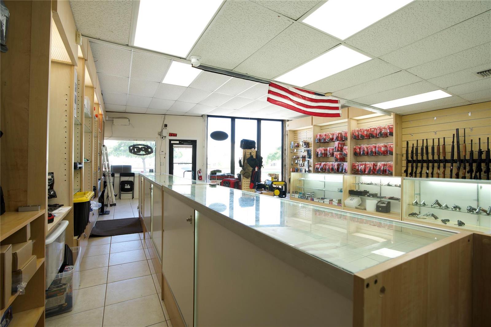 A retail store interior features glass display cases, wall-mounted racks for firearms, and a ceiling-mounted flag — 4581 N Dixie Highway, Deerfield Beach FL 33064, DEERFIELD BEACH, FL