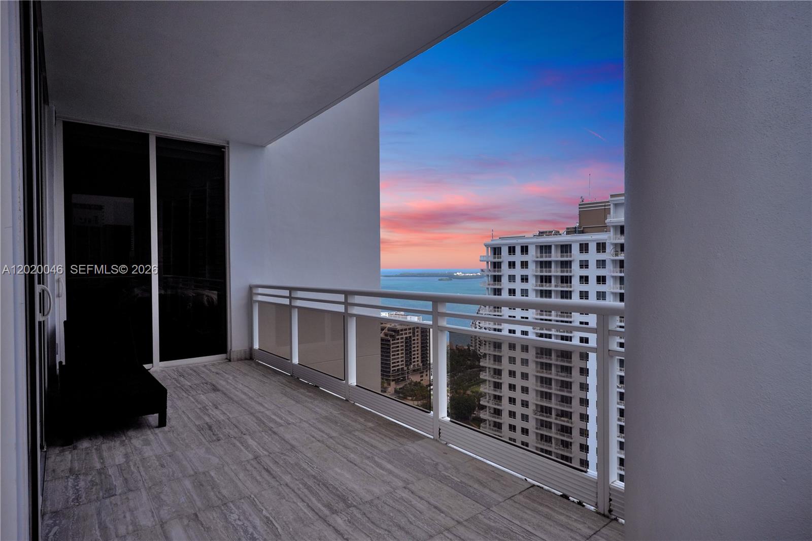A balcony with light-colored tile flooring overlooks a bay and neighboring high-rise buildings at sunset — 901 Brickell Key Blvd # 3204, Miami FL 33131, MIAMI, FL