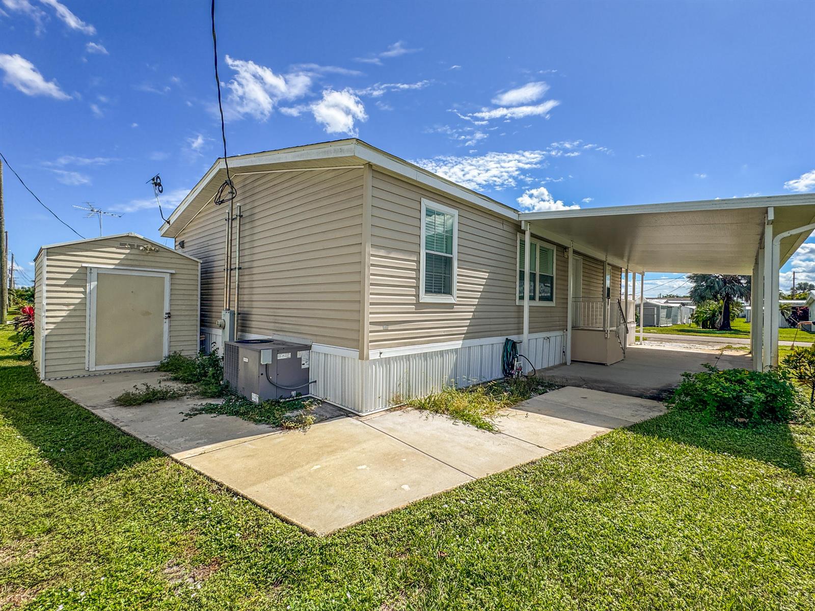 A tan mobile home with a carport, attached shed, and concrete patio under a blue sky — 4377 Mission Bell Drive, Boynton Beach FL 33436, BOYNTON BEACH, FL