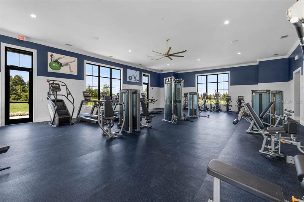 A fitness center features various exercise machines, dark rubber flooring, and large windows with outdoor views — 7110 SANDHILL CRANE WAY UNIT 1, ST CLOUD, FL