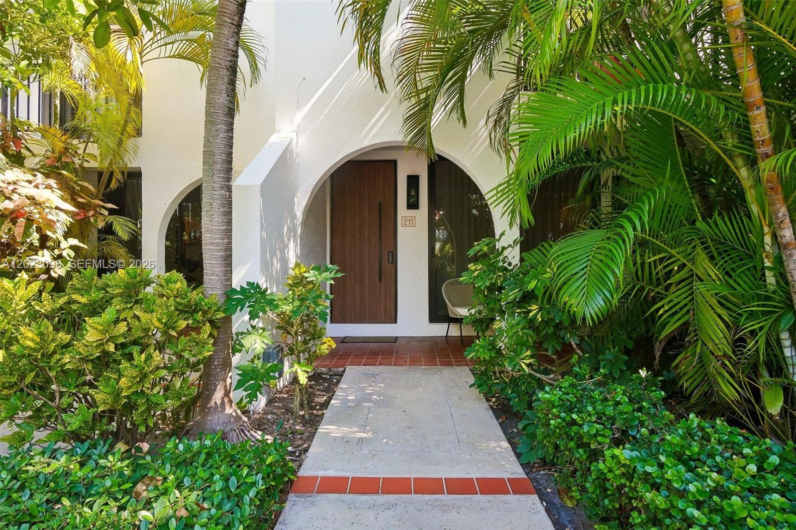 A concrete walkway leads to the arched entryway of a white building with a wooden door — 425 Grapetree Dr # 211, Key Biscayne FL 33149, KEY BISCAYNE, FL