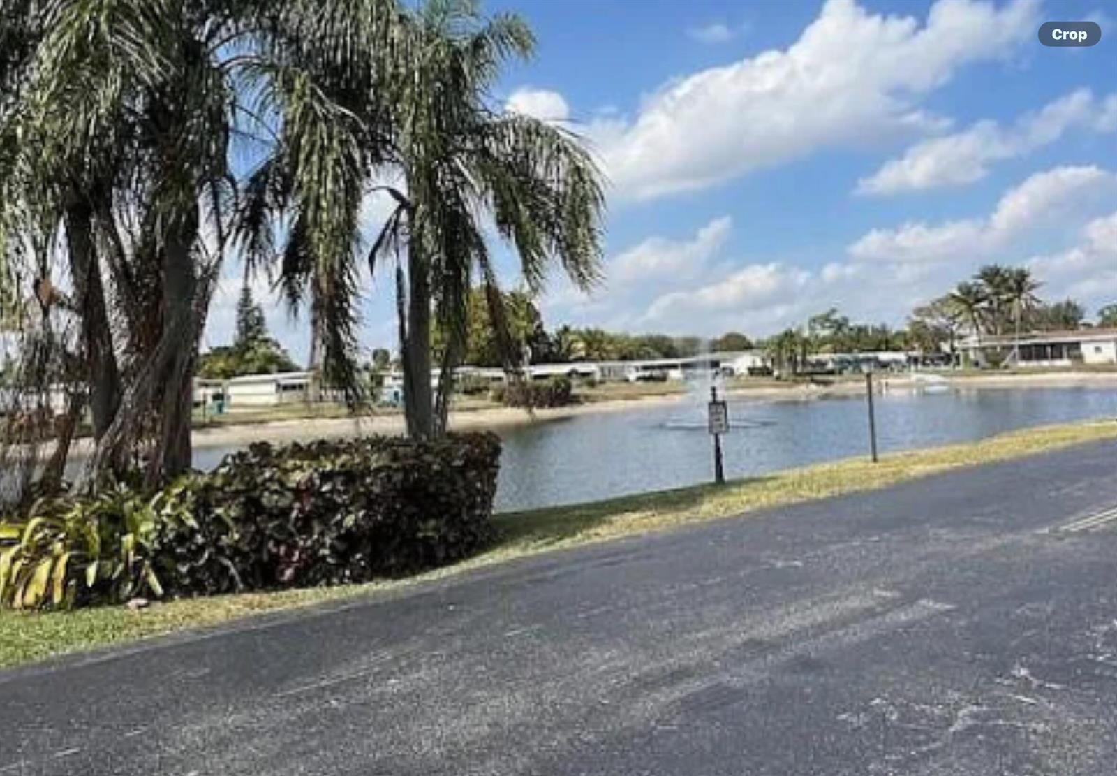A view of a pond with a fountain, palm trees, and mobile homes in the background — 8628 Duke Court E, Boynton Beach FL 33436, BOYNTON BEACH, FL