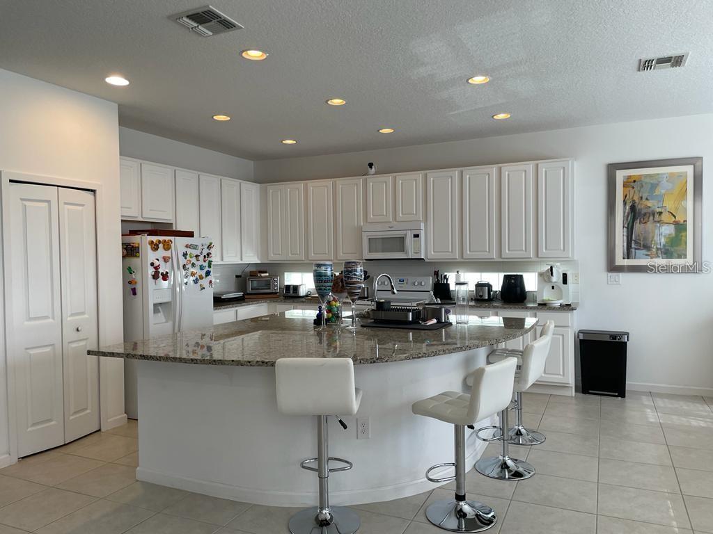 A kitchen features white cabinetry, granite countertops, a curved island with bar stools, and tile flooring — 3850 OAKVILLE AVENUE, KISSIMMEE, FL
