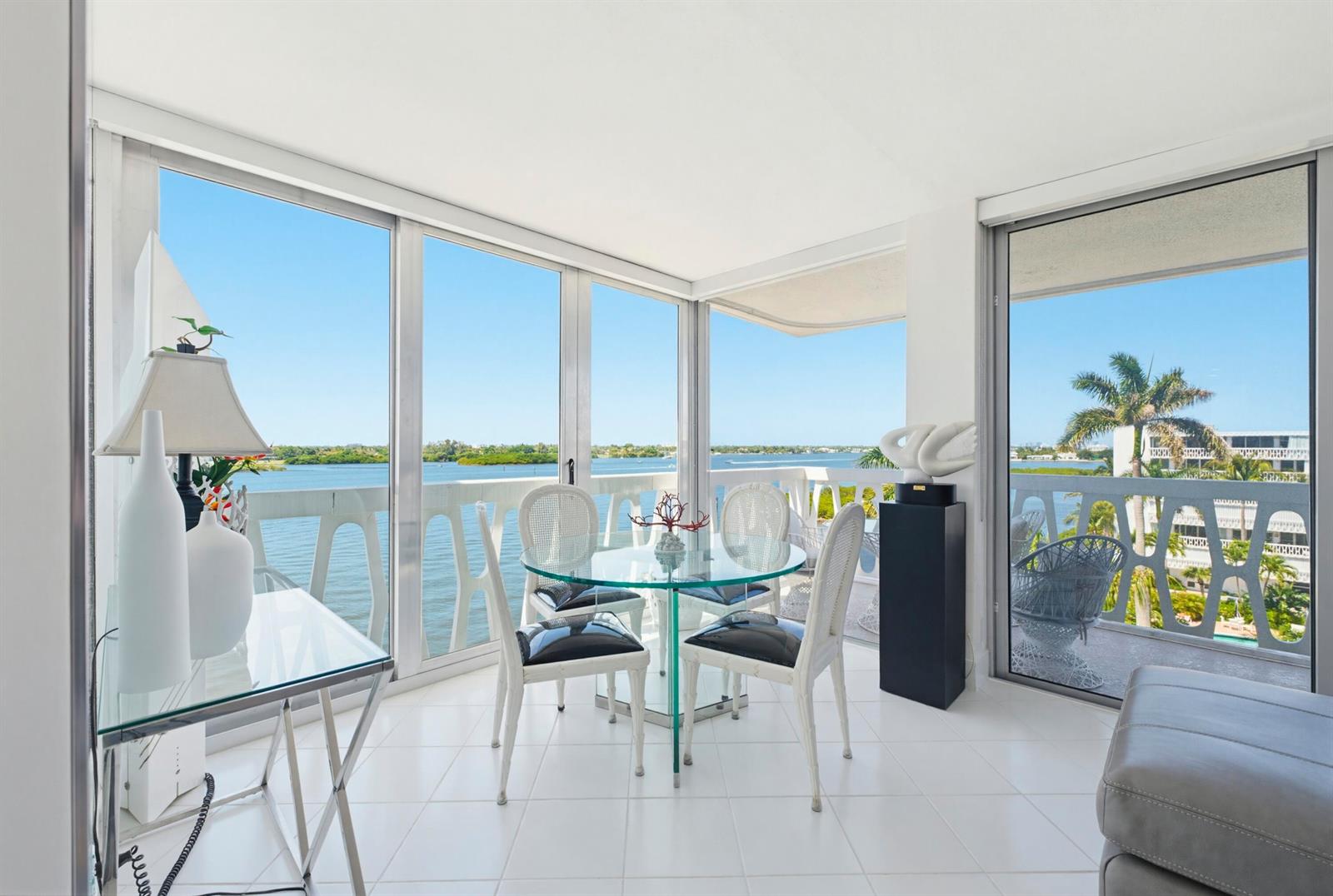 A bright dining area features a glass table, white chairs, and floor-to-ceiling windows overlooking water — 2505 S Ocean Boulevard, Palm Beach FL 33480, PALM BEACH, FL