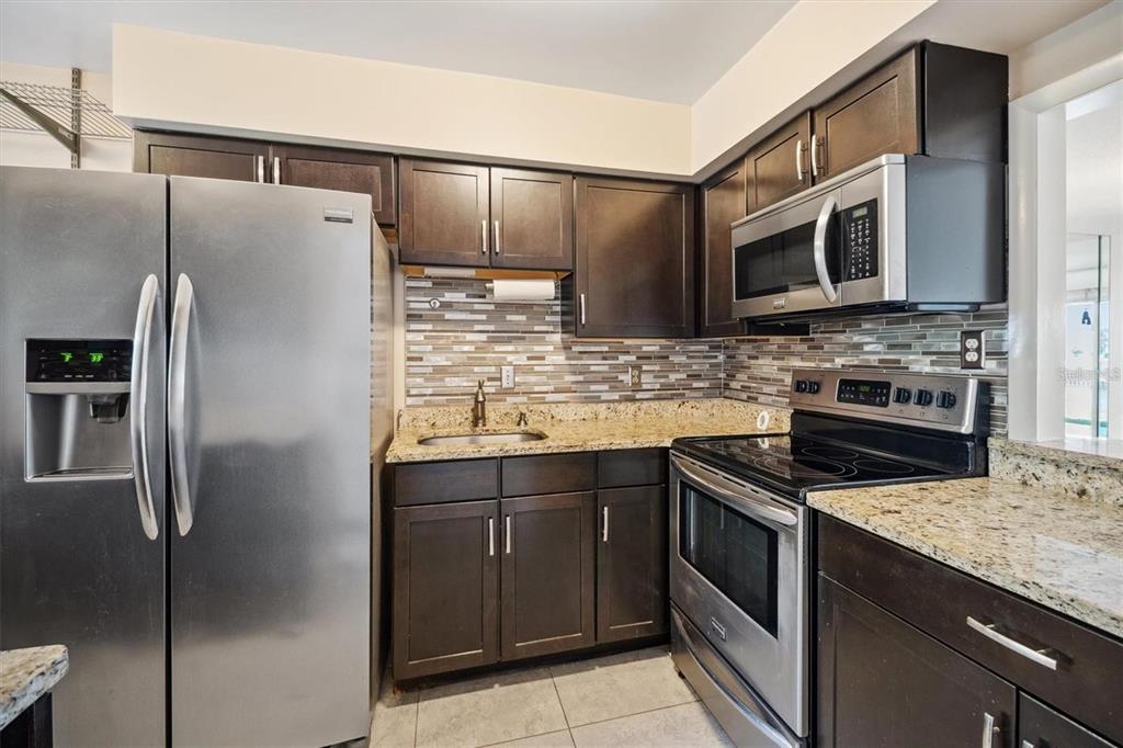 A kitchen featuring dark wood cabinets, granite countertops, stainless steel appliances, and a tiled backsplash — 44 CLUB HOUSE DRIVE UNIT 105, PALM COAST, FL