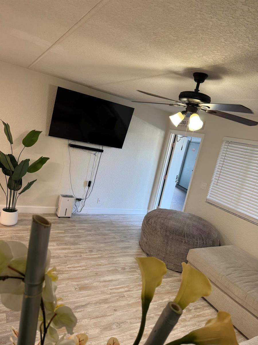 A living room features a wall-mounted television, a ceiling fan, and light wood-look flooring — 2000 Springdale Boulevard # 108, Palm Springs FL 33461, PALM SPRINGS, FL