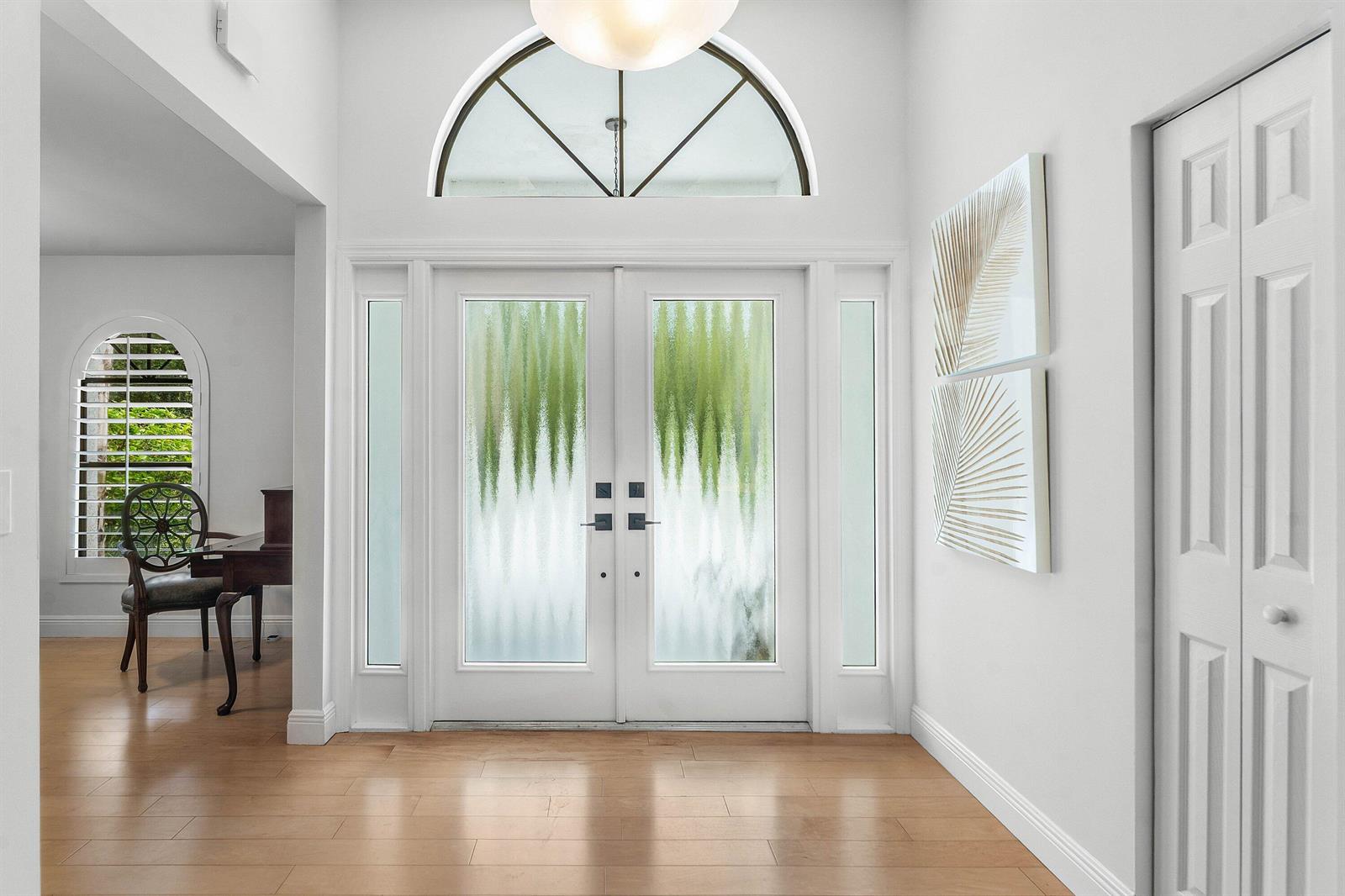 A bright entryway features double glass doors, light wood flooring, and a white arched transom window — 1488 SE Colony Way, Jupiter FL 33478, JUPITER, FL