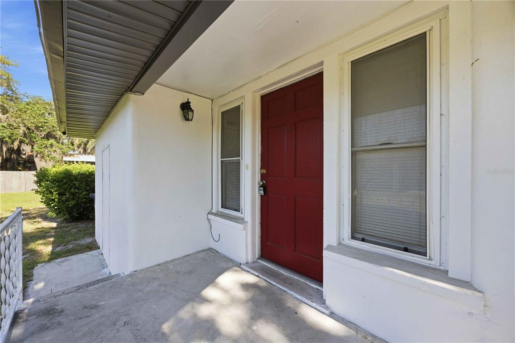 A covered concrete porch features a red front door and a window with blinds — 1997 26TH STREET NW, WINTER HAVEN, FL