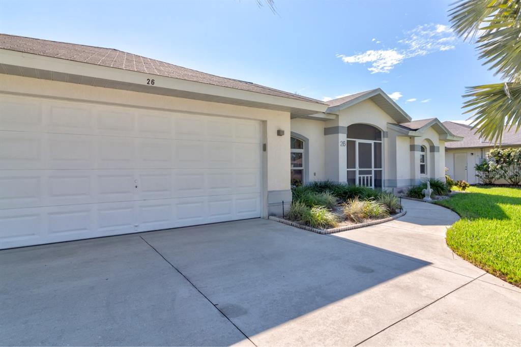 A single-story house exterior features a white garage door, a screened entryway, and landscaping — 26 LONG MEADOW LANE, ROTONDA WEST, FL