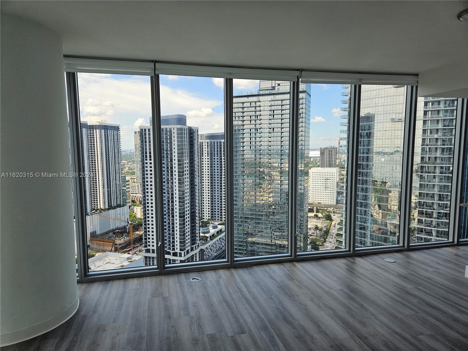 An empty room with wood-look flooring features floor-to-ceiling windows overlooking a dense urban skyline — 888 Biscayne Blvd # 4001, Miami FL 33132, MIAMI, FL