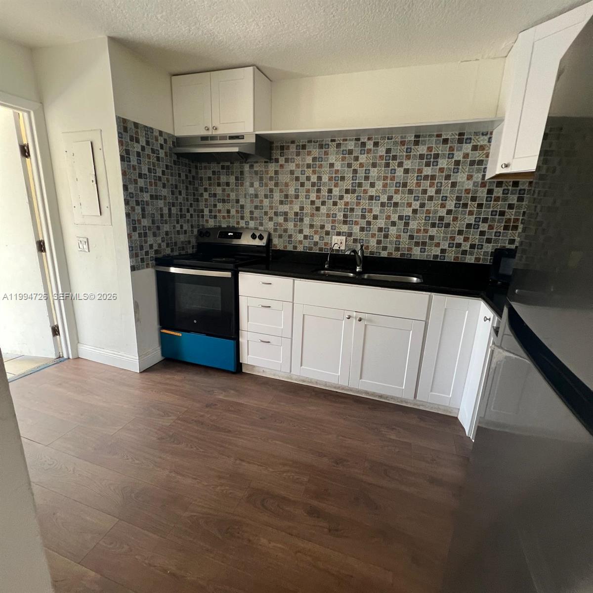 A kitchen features white cabinets, a black countertop, a mosaic tile backsplash, and wood flooring — 109 SW 1st Ave, Hallandale Beach FL 33009, HALLANDALE BEACH, FL