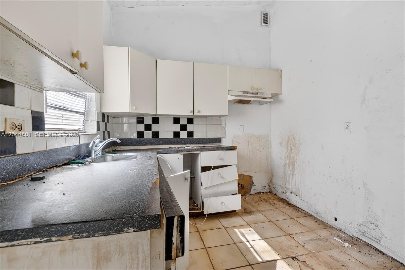 A dilapidated kitchen features white cabinets, a dark countertop, and stained, damaged walls and flooring — MIAMI, FL