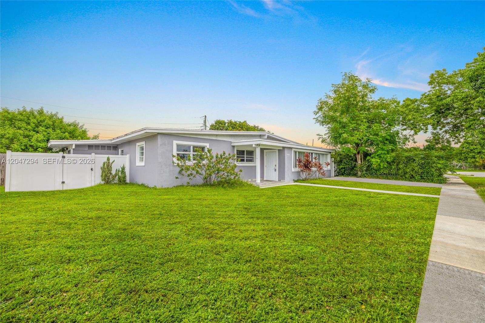 A single-story gray house with a white fence and a green lawn under a blue sky — 18100 NW 7th Pl, Miami Gardens FL 33169, MIAMI GARDENS, FL