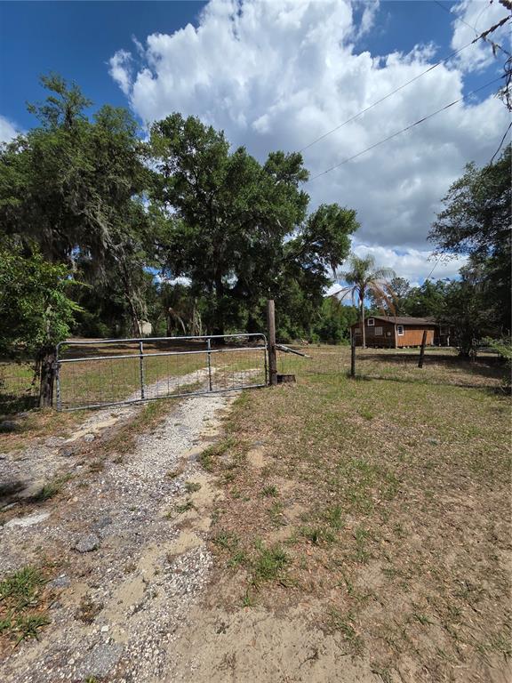 A metal gate opens to a gravel driveway leading toward a small brown house — 27440 WINCHESTER DRIVE, BROOKSVILLE, FL