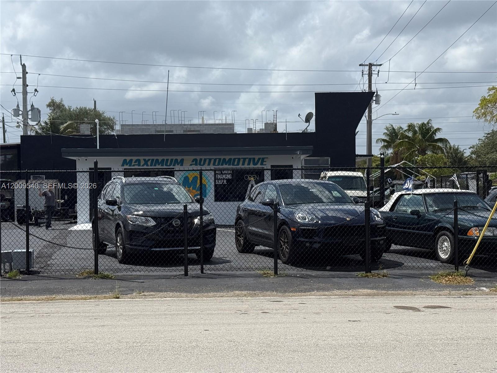 A black building labeled Maximum Automotive sits behind a chain-link fence with parked cars — 1822 N Dixie Hwy, Hollywood FL 33020, HOLLYWOOD, FL