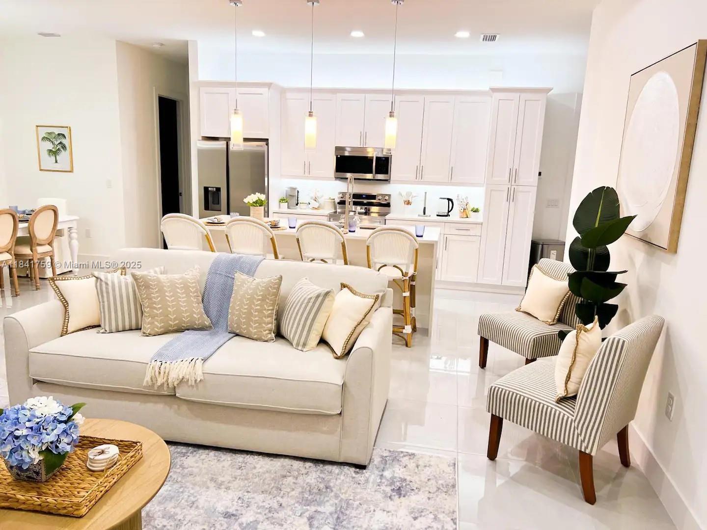 A bright open-concept living room and kitchen featuring white cabinetry, neutral furniture, and tile flooring — 404 NW 1st ST, Cape Coral FL 33993, CAPE CORAL, FL
