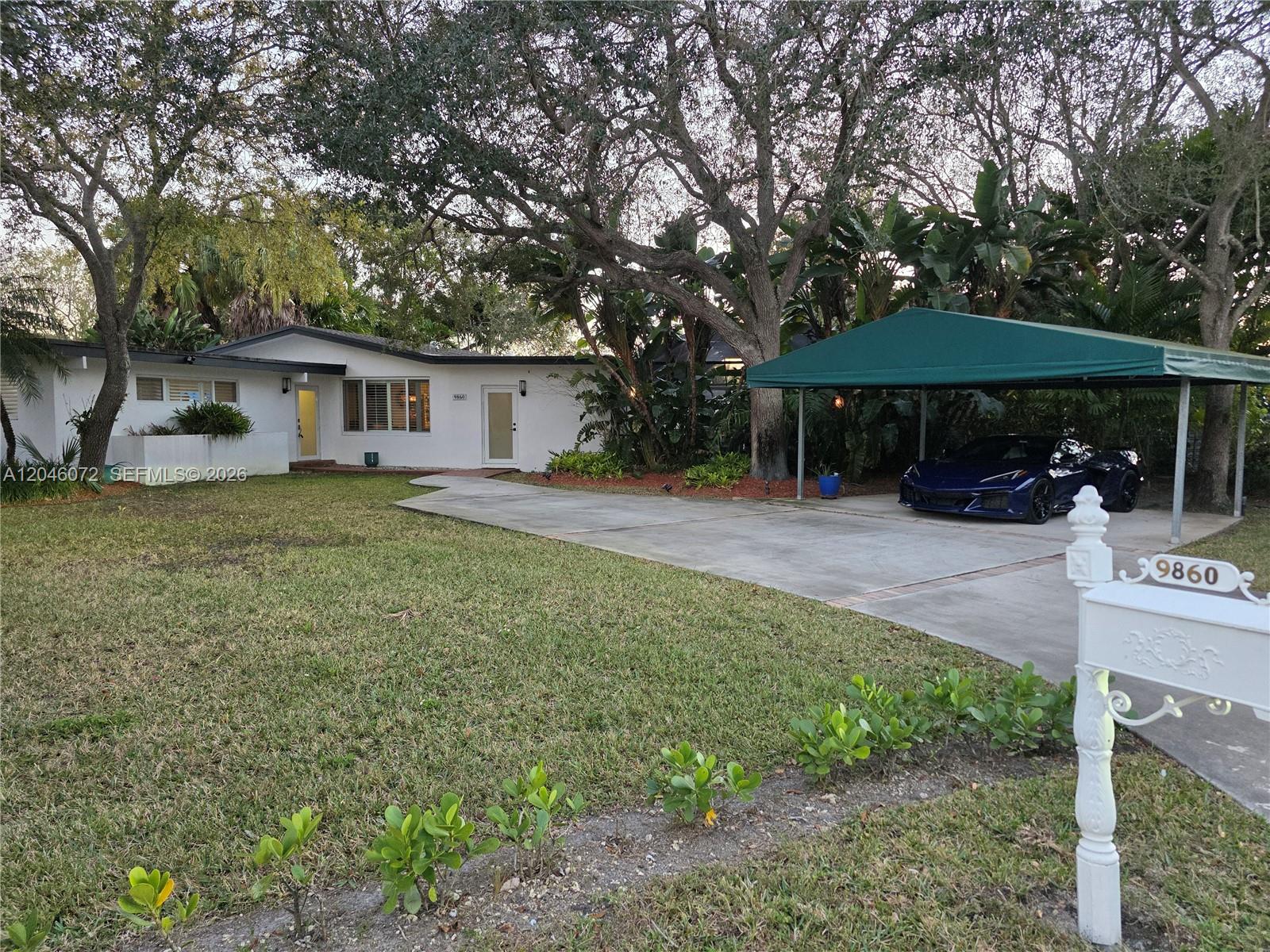 A white single-story house features a green carport sheltering a dark blue sports car — 9860 SW 140th St, Miami FL 33176, MIAMI, FL