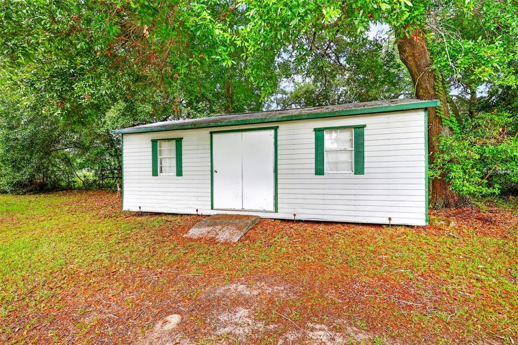 A white storage shed with green trim sits on a grassy lot under large trees — 15900 SE 92ND TERRACE, SUMMERFIELD, FL