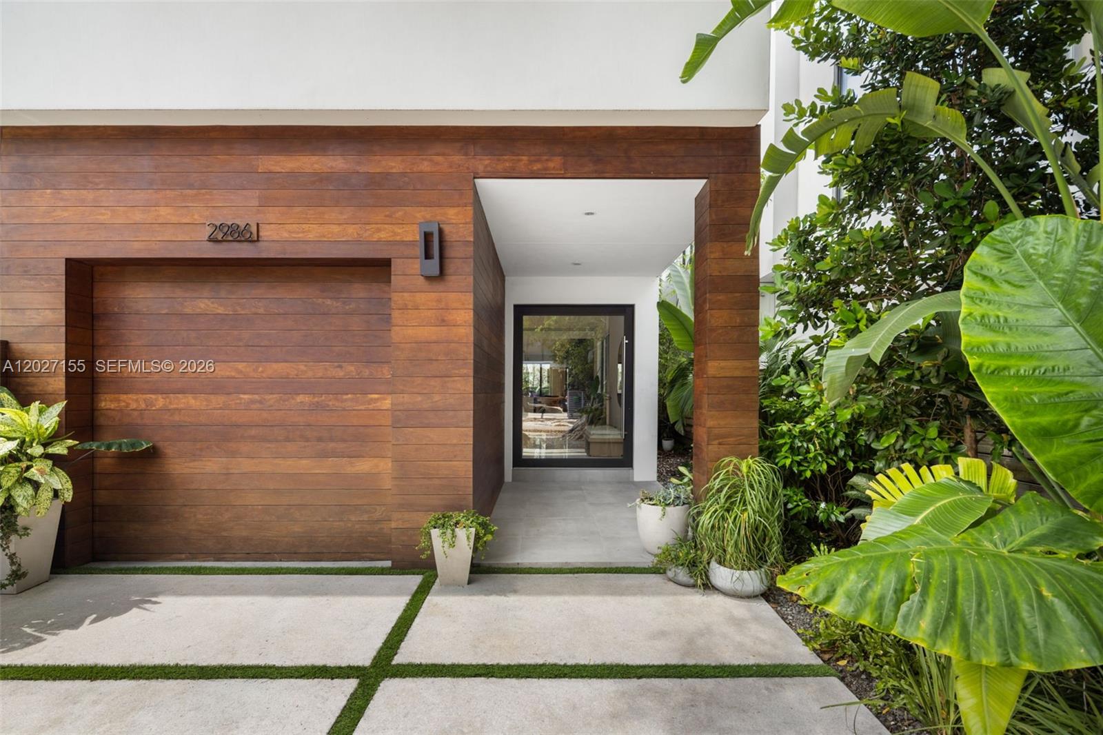 A modern home exterior features a wood-paneled garage, a glass front door, and lush landscaping — 2986 Coconut Ave, Coconut Grove FL 33133, COCONUT GROVE, FL