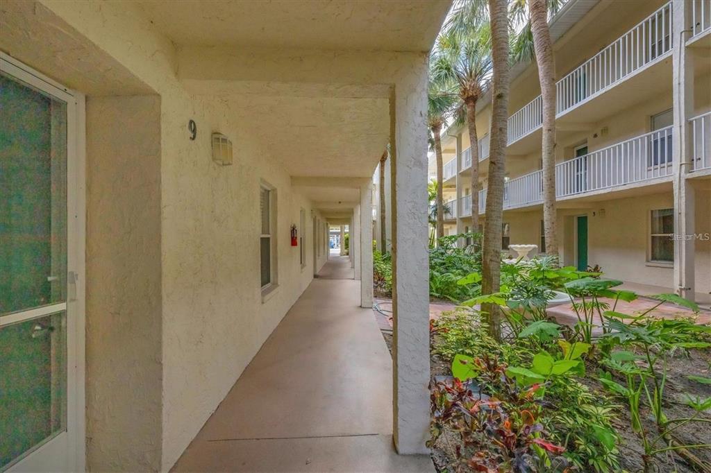 An outdoor covered walkway leads past apartment units with a garden area to the right — 7294 CLOISTER DRIVE UNIT 9, SARASOTA, FL
