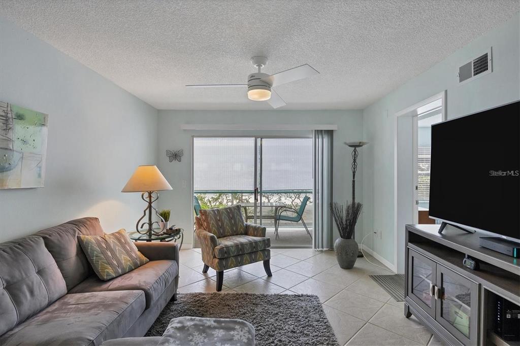 A living room features a grey sofa, patterned armchair, ceiling fan, and sliding glass balcony doors — 7294 CLOISTER DRIVE UNIT 9, SARASOTA, FL