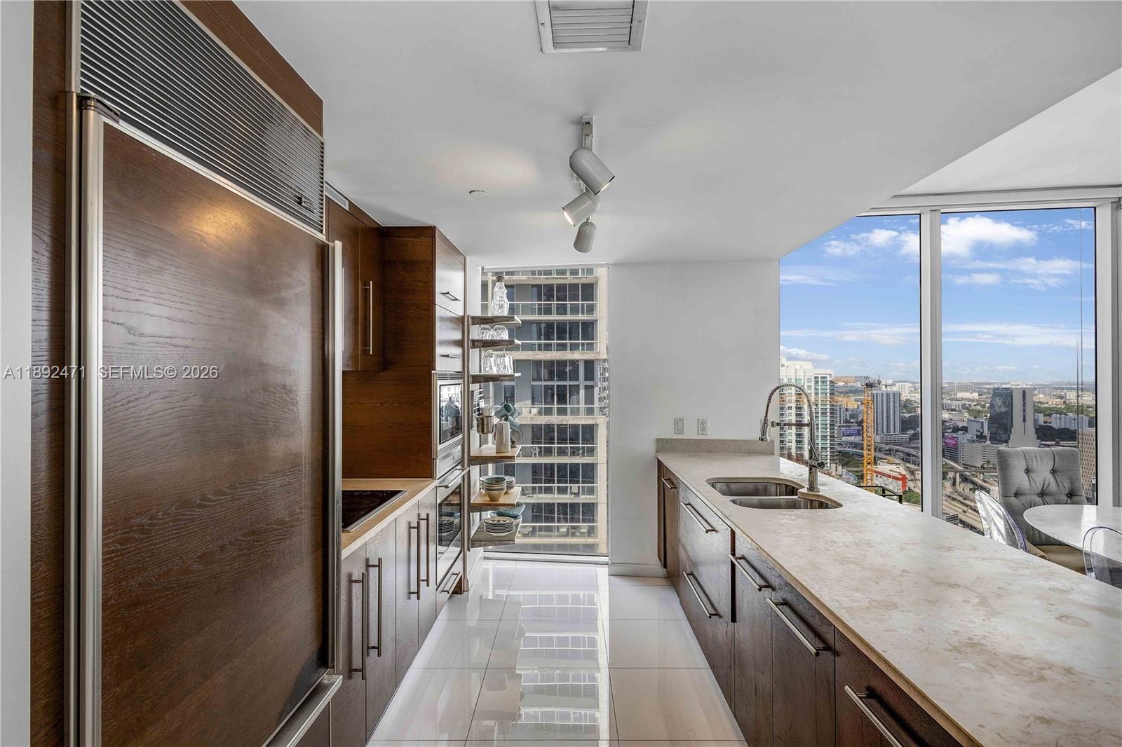 A modern galley kitchen features wood cabinetry, stone countertops, and floor-to-ceiling windows with city views — 475 Brickell Ave # 4015, Miami FL 33131, MIAMI, FL