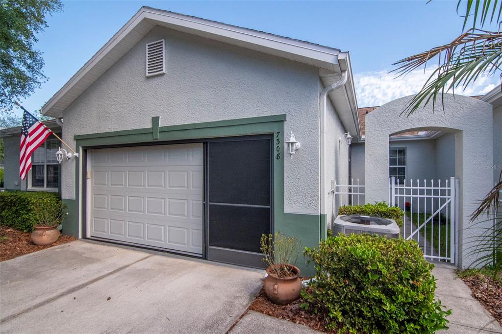 A house exterior features a garage with a screen door, a white gate, and landscaping — 7308 CLEOPATRA DRIVE, LAND O LAKES, FL
