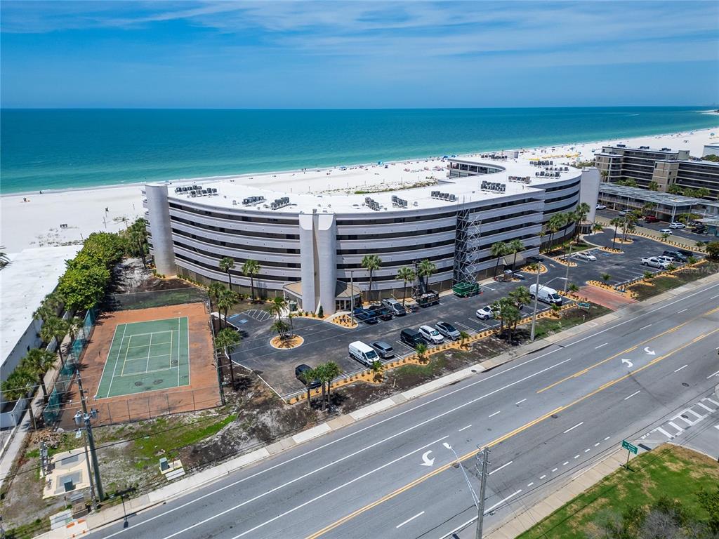 An aerial view of a curved beachfront condominium building with a tennis court and parking — 4450 GULF BOULEVARD UNIT 113, ST PETE BEACH, FL
