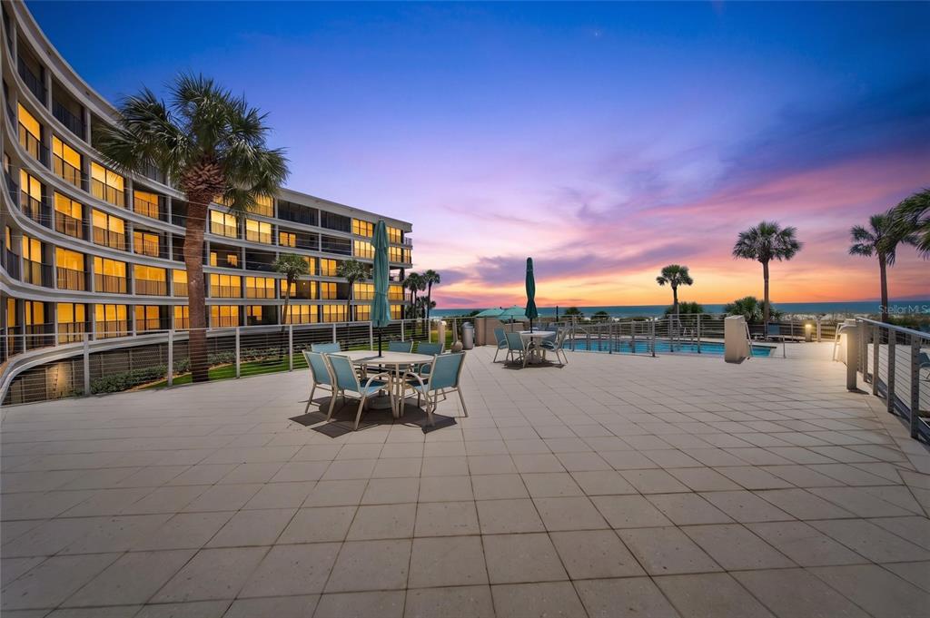 A patio with tables and chairs overlooks a swimming pool and the ocean at sunset — 4450 GULF BOULEVARD UNIT 113, ST PETE BEACH, FL