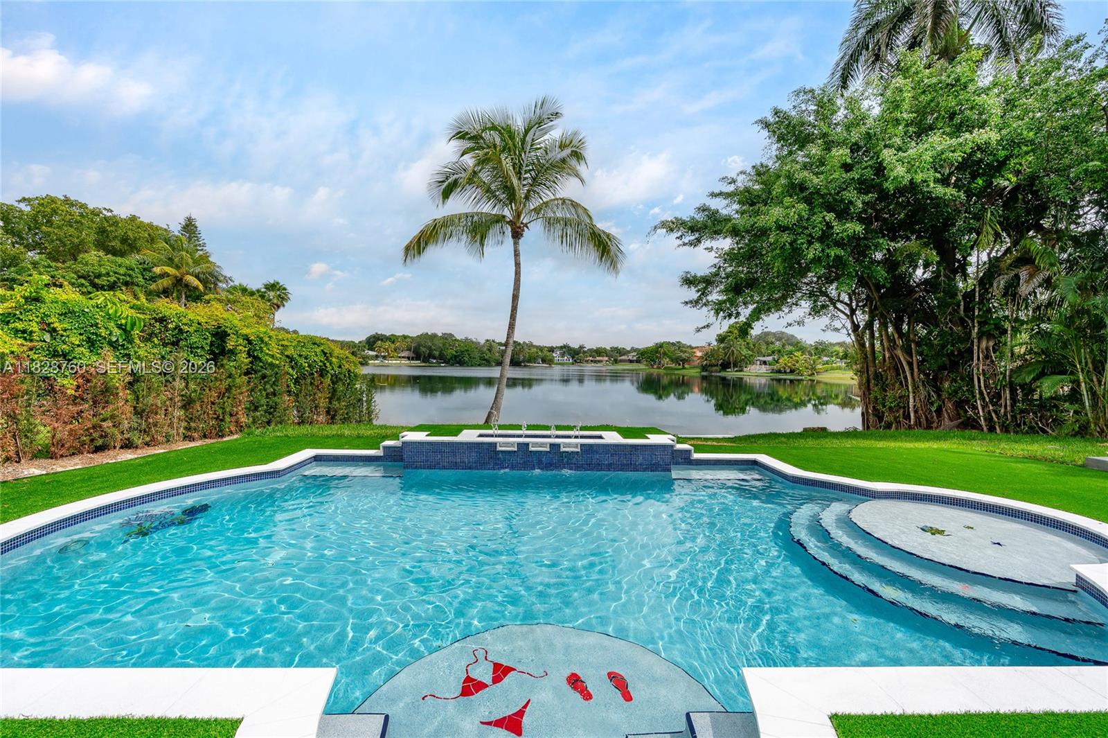 A backyard swimming pool with a water feature overlooks a lake and palm trees — 7101 E Troon Cir, Miami Lakes FL 33014, MIAMI LAKES, FL