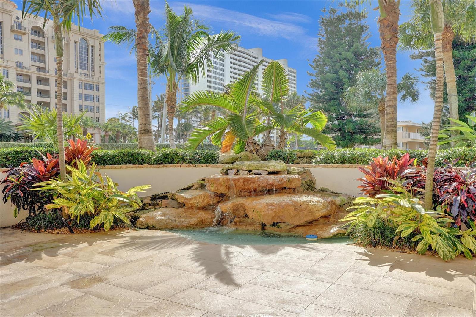 A stone waterfall feature sits in a landscaped patio area with palm trees and buildings — 2501 S Ocean Boulevard # 1030, Boca Raton FL 33432, BOCA RATON, FL