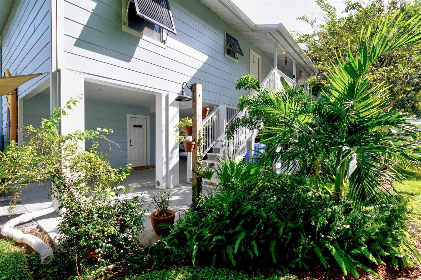 A light blue elevated house features a ground-level patio, white stairs, and lush tropical landscaping — FELLSMERE, FL