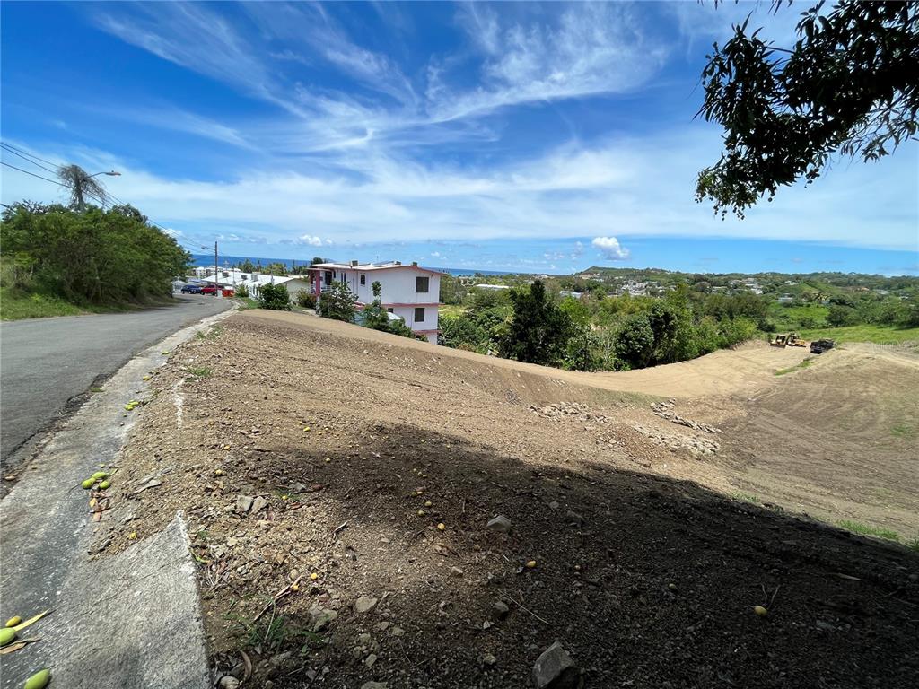 A sloped, cleared dirt lot sits beside a paved road with a distant ocean view — SIX LOTS FOR SALES PRICE START $170K, RINCON, PR