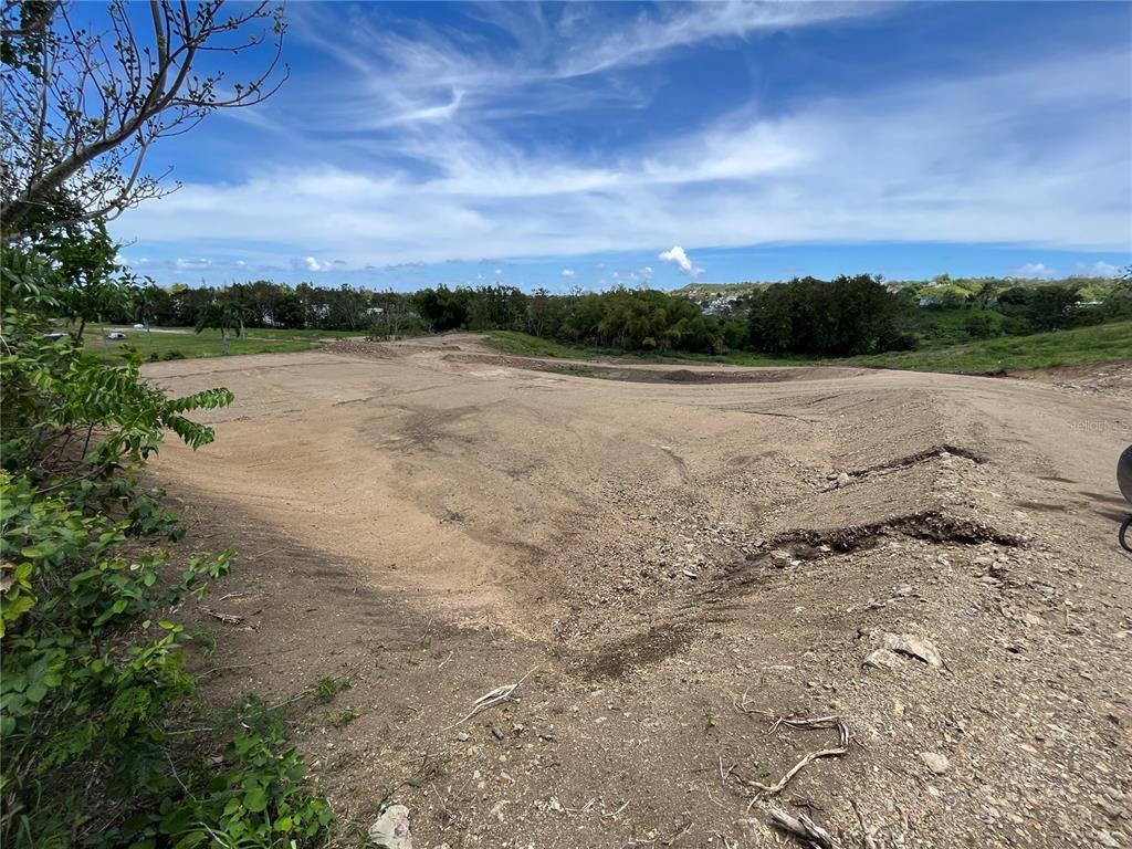 A large, cleared dirt lot sits under a blue sky with distant trees and hills — SIX LOTS FOR SALES PRICE START $170K, RINCON, PR