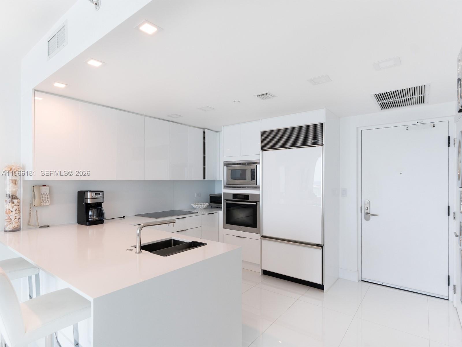 A modern white kitchen features a central island with a sink and built-in appliances — 6799 Collins Ave # 704, Miami Beach FL 33141, MIAMI BEACH, FL